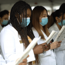 A group of students in white lab coats and masks standing and reading a ceremony program